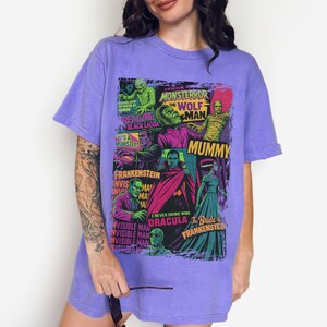 May include: A lilac t-shirt with a vibrant graphic of classic Universal Monsters film posters. The design includes images of Frankenstein, Dracula, The Mummy, and The Wolfman. The t-shirt has a loose fit.
