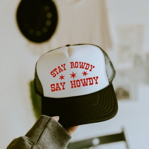 May include: White trucker hat with a black mesh back. The hat has red text that says "Stay Rowdy Say Howdy" with three red stars.