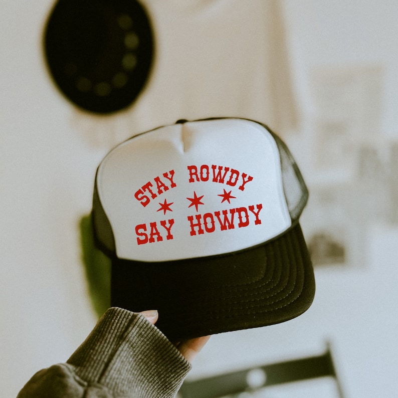 May include: White trucker hat with a black mesh back. The hat has red text that says "Stay Rowdy Say Howdy" with three red stars.