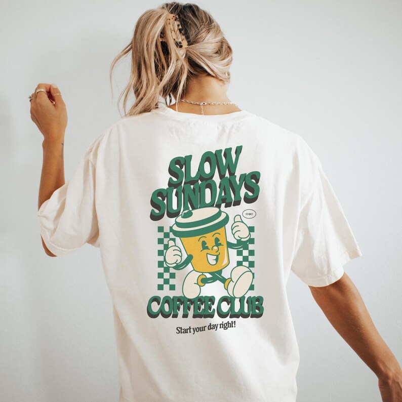 May include: Cream-colored t-shirt with a retro-style graphic on the back. The graphic features a smiling coffee cup character with the words "SLOW SUNDAYS COFFEE CLUB" in green. The phrase "Start your day right!" is printed below.