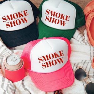 May include: Three trucker hats in black, green, and pink, each with a white front panel. The hats feature the text "SMOKE SHOW" in red. Accessories include a pink drinks cooler, sunglasses, and a can of soda.