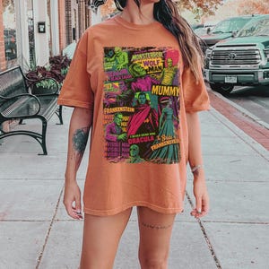 May include: A rust-coloured t-shirt with a retro graphic design showcasing iconic monster movie characters, including Frankenstein, Dracula, and the Mummy. The design features the text "Monster Terror", "Wolf Man", and "Mummy".