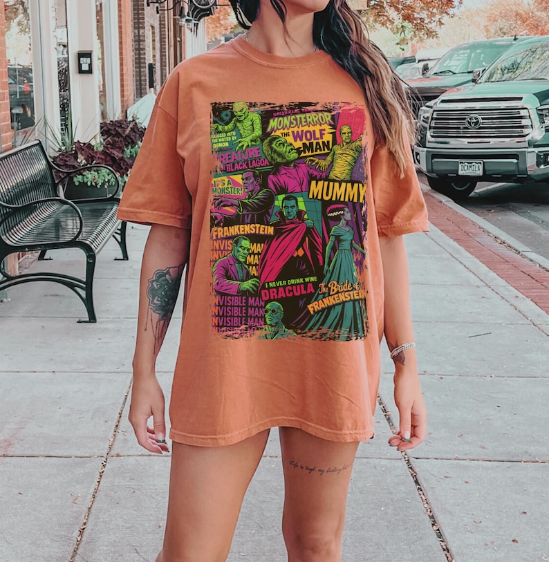 May include: A rust-coloured t-shirt with a retro graphic design showcasing iconic monster movie characters, including Frankenstein, Dracula, and the Mummy. The design features the text "Monster Terror", "Wolf Man", and "Mummy".