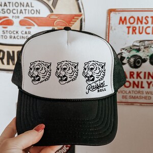 May include: A black and white trucker hat with a white mesh back. The front of the hat features three black and white tiger head illustrations and the text "Raisin Hell".