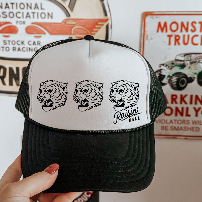 May include: A black and white trucker hat with a white mesh back. The front of the hat features three black and white tiger head illustrations and the text "Raisin Hell".