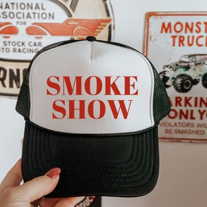 May include: A black and white trucker hat with the text "SMOKE SHOW" in red. The hat features a mesh back and a curved peak. The hat is held up in front of a wall with vintage signs.