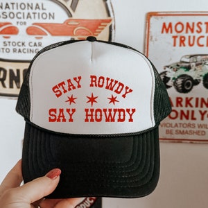 May include: A white and black trucker hat with a red embroidered design that says "Stay Rowdy *** Say Howdy".