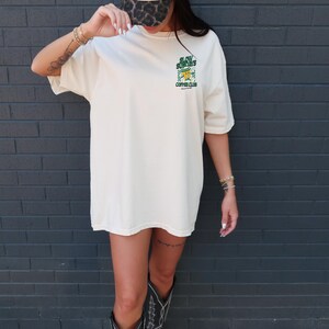 May include: Cream-colored oversized t-shirt with a green and yellow graphic that reads "Slow Sundays Coffee Club." The shirt is paired with a leopard print baseball cap and black cowboy boots. The background is a dark gray brick wall.