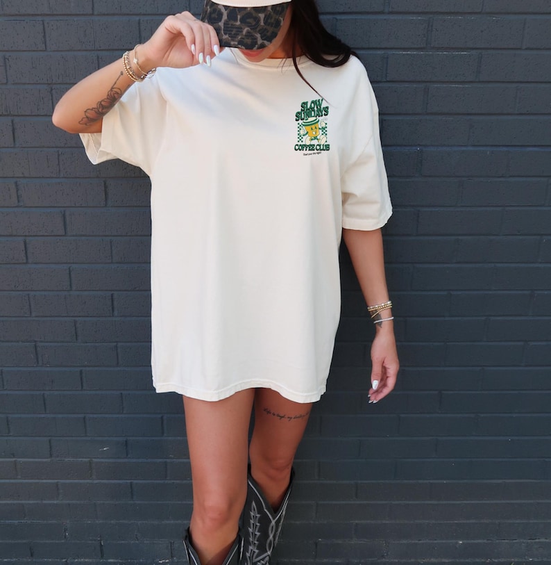 May include: Cream-colored oversized t-shirt with a green and yellow graphic that reads "Slow Sundays Coffee Club." The shirt is paired with a leopard print baseball cap and black cowboy boots. The background is a dark gray brick wall.