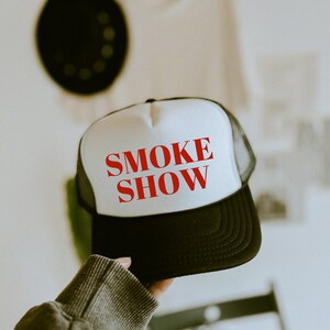 May include: A black and white trucker hat with the words "SMOKE SHOW" in red. The hat features a white front panel, a black brim, and a black mesh back. The hat is held up by a person.