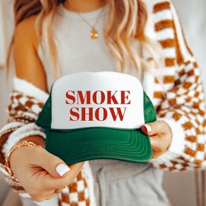 May include: A green and white trucker hat with the words "SMOKE SHOW" in red. The hat features a white front panel and a green mesh back. The hat is being held up.