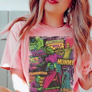 May include: Pale pink t-shirt with a vibrant graphic showcasing classic Universal Monsters film posters. The design includes images of Frankenstein, Dracula, the Mummy, and the Wolf Man, with text that reads "MONSTERROR" and "DRACULA".