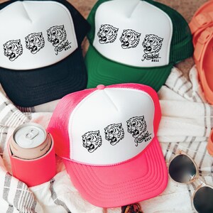 May include: Three pink, green, and black trucker hats with white mesh backs. Each hat features a white graphic of a tiger's face and the text "Raisin' Hell".