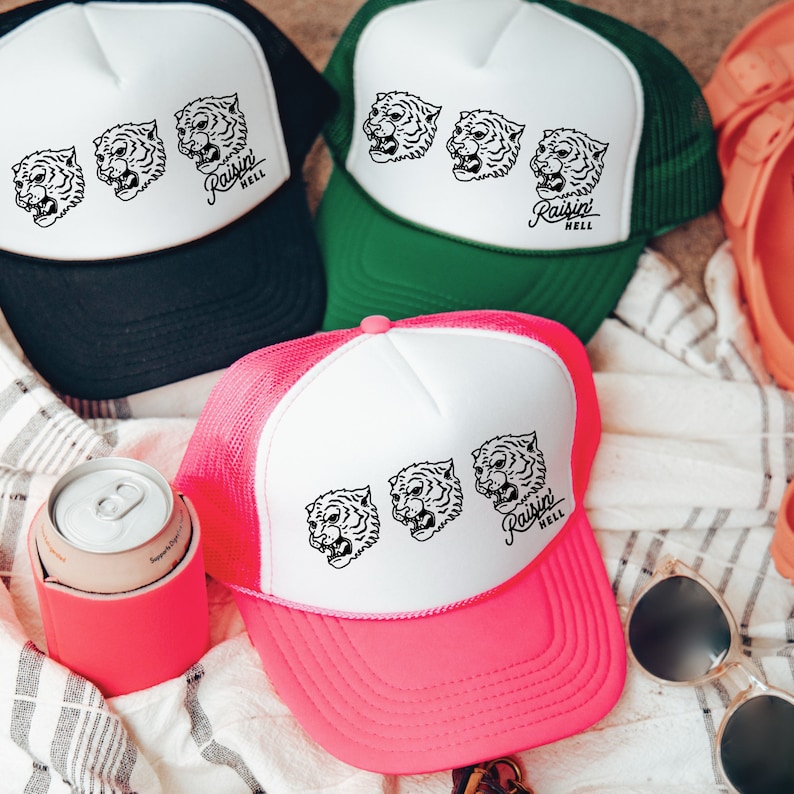 May include: Three pink, green, and black trucker hats with white mesh backs. Each hat features a white graphic of a tiger's face and the text "Raisin' Hell".