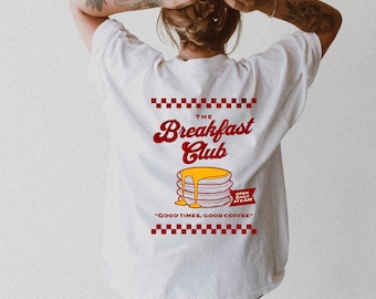 May include: White t-shirt with a red and yellow graphic design on the back. The design features the words "The Breakfast Club" above a stack of pancakes with syrup, and the phrase "Good Times, Good Coffee."