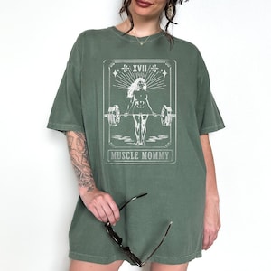 May include: Oversized green t-shirt featuring a white graphic of a woman lifting weights. The graphic is in the style of a tarot card and says "Muscle Mommy" at the bottom.