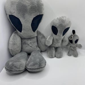 以下が含まれることがあります： さまざまなサイズの3つの灰色のぬいぐるみエイリアンの玩具。最大のエイリアンは座っており、大きな黒い目の形と胸にステッチされたXがあります。他の2つのエイリアンは立っており、1つはキーホルダーが付いています。すべて柔らかくふわふわした質感です。