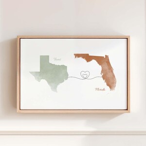 May include: A framed wall art print featuring a sage green watercolour map of Texas and a terracotta watercolour map of Florida. A thin black line connects the two states with a small heart outline in the centre. The words Texas and Florida are printed.