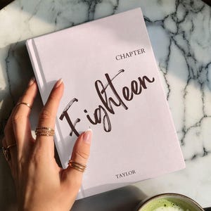 May include: A book titled "Eighteen" with the author's name "Taylor" is held by a hand adorned with gold rings and bracelets. A cup of green matcha latte sits nearby on a marble surface.
