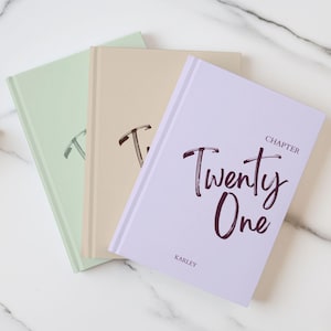 May include: Three hardcover journals in pastel shades of light green, beige, and lavender. The lavender journal features the dark purple text "CHAPTER Twenty One" and the name "KARLEY" printed on the front cover in a stylized script font.