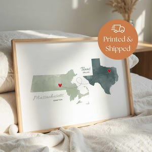 May include: A framed watercolor map print featuring the states of Massachusetts and Texas, with red heart markers indicating locations. The text "Massachusetts Grafton" and "Texas Dallas" are included. The print has a light wooden frame.