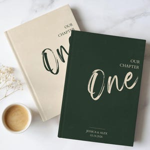 May include: Two hardback books with matte covers, one in cream and one in dark green. Both feature the text "OUR CHAPTER" above the word "One" in a brushstroke font. The green book includes the names "JESSICA & ALEX" and "03.18.2026" at the bottom.
