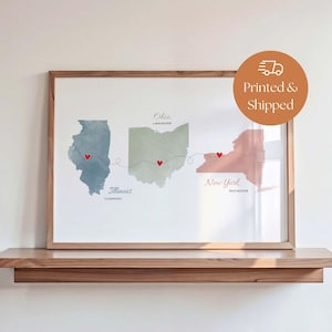 May include: Framed watercolor print featuring the states of Illinois, Ohio, and New York. Each state is depicted in a different color, with red heart markers and dotted lines connecting the cities of Clermont, Lancaster, and Rochester. The print is displayed on a wooden shelf.