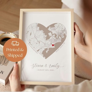 May include: A framed print featuring a heart-shaped map of Saint John, NB, with a small red heart icon. The text reads Our Engagement, Steven & Emily, and August 16th, 2024. The print is housed in a light-coloured wooden frame with a white border.