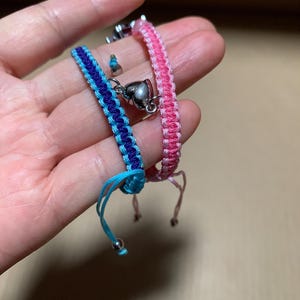 Puede incluir: Dos pulseras de la amistad con dijes en forma de coraz&oacute;n. Una pulsera es rosa y la otra es azul y negra.