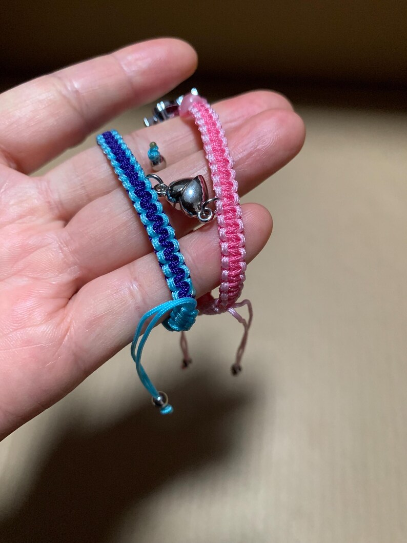 Puede incluir: Dos pulseras de la amistad con dijes en forma de coraz&oacute;n. Una pulsera es rosa y la otra es azul y negra.