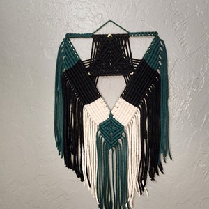 Puede incluir: Un tapiz de macramé de pared de color negro, blanco y verde con un diseño geométrico. El tapiz tiene una forma triangular con flecos en la parte inferior.