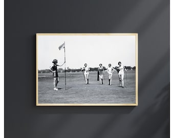 May include: A black and white framed photograph depicting five individuals on a golf course. One person stands by a flagstick while four others walk across the grass carrying golf clubs. The scene captures a vintage outdoor sporting activity.