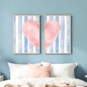 Op de afbeelding: Twee rechthoekige wandkunstprints in lichte houten lijsten. Elke print heeft een achtergrond met verticale blauwe en witte strepen en de helft van een groot, zachtroze aquarelhart, die samen een compleet hart vormen.