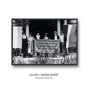 May include: A black and white photograph featuring six individuals standing in a row holding a banner that reads: NO SELF RESPECTING WOMAN SHOULD WISH OR WORK FOR THE SUCCESS OF A PARTY THAT IGNORES HER SEX. SUSAN B. ANTHONY 1872.
