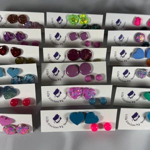 May include: A collection of stud earrings displayed on white cards, featuring various shapes including hearts, circles, and squares in diverse colours like pink, blue, green, and purple, with some designs incorporating glitter and iridescent finishes.