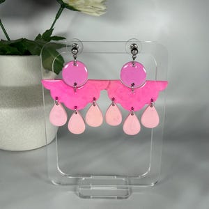 May include: A pair of dangle earrings featuring a circular top piece in bright pink, a scalloped semi-circular middle section in hot pink, and three dangling teardrop-shaped charms in light pink, all connected by silver-toned metal jump rings.