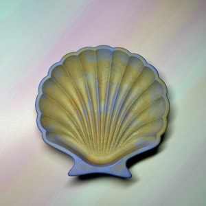 May include: A decorative seashell-shaped dish with a scalloped edge. The dish has a light blue rim and a golden interior with radial lines. The dish is designed to resemble a scallop shell and is suitable for holding small items.