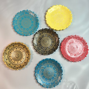 Puede incluir: Cinco platos decorativos con diseños de mandalas. Los platos son de varios colores, incluyendo amarillo, azul, turquesa, negro y rojo. Cada plato tiene un diseño de mandala dorado.