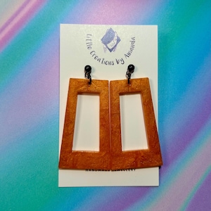 May include: A pair of gold-coloured rectangular earrings with black posts. The earrings are made of resin and have a textured surface. The earrings are handmade and are labelled "little creations by Amanda".