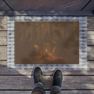 Puede incluir: Un felpudo rectangular con un centro marrón oscuro que presenta una tenue silueta naranja brillante de un castillo y árboles, rodeado por un borde ancho a cuadros de color gris claro y blanco, sobre una superficie de madera.