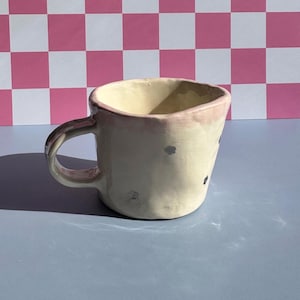 May include: A white ceramic mug with a pink rim and black speckles. The mug has a handle and is sitting on a light blue surface.