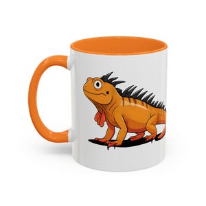 May include: A white ceramic mug with an orange rim and handle. The mug features a cartoon illustration of an orange iguana with black stripes and red accents.