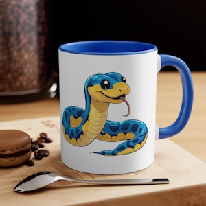 May include: A white ceramic mug with a blue rim and handle. The mug features a cartoon illustration of a blue and yellow snake with big eyes and a tongue sticking out.