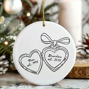 May include: A white ceramic Christmas ornament with a gold string for hanging. The ornament features a black line drawing of two heart-shaped lockets, one labeled "Mom est. 1995" and the other "Grandma est. 2025". A bow is drawn above the lockets.