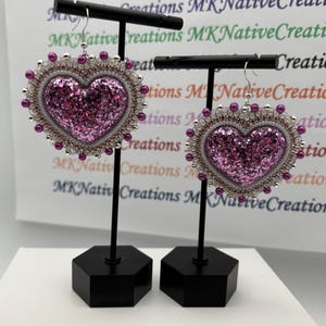 May include: Two heart-shaped earrings with a glittery pink center, surrounded by white and pink beads. The earrings are displayed on black stands against a background with the text "MK Native Creations".
