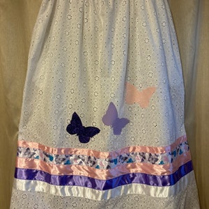 May include: A white skirt with a floral pattern and three butterfly appliques. The skirt has a purple, pink, and white ribbon trim.