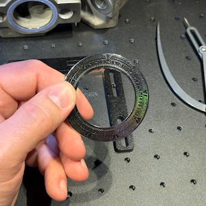 Puede incluir: Una mano sosteniendo un objeto metálico circular oscuro con marcas grabadas. El objeto tiene un centro transparente y colores iridiscentes. Un compás plateado y otras piezas mecánicas están en el fondo.