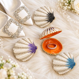 May include: White satin high-heeled shoes with large crystal buckles and several seashell-shaped trinket boxes. The boxes feature white ridges with crystal accents and colored tips in black, purple, and blue. One open box contains a diamond ring.