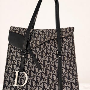 May include: A black and beige tote bag featuring a repeating monogram pattern. The bag has black leather straps, a black leather flap closure, and a silver-toned metal letter D charm hanging from the front strap. The design includes a structured shape.