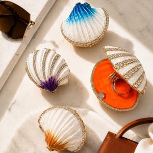 May include: Four shell-shaped jewelry boxes with gold-tone metal edges and clear crystal accents. One box is open, revealing a bright orange interior and a ring. The others are closed, featuring white enamel with blue, purple, or tan gradients.